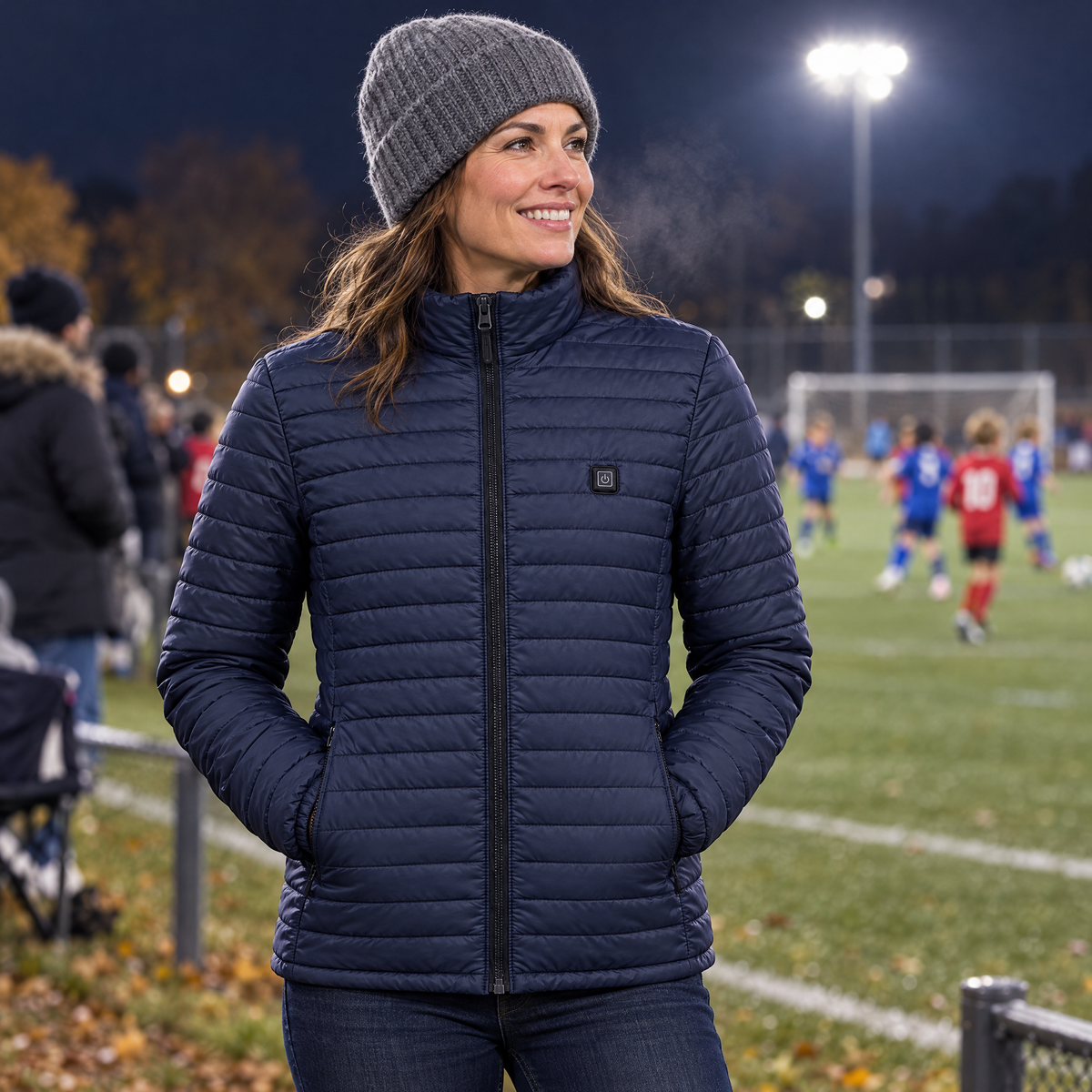 Mom at a youth soccer game under the lights, wearing the Basecamp women's heated jacket in navy, hands in the heated pockets