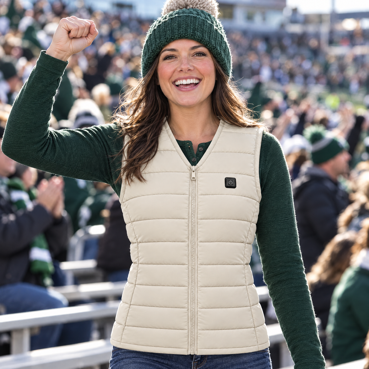 Woman cheering at a college football game wearing the Ridgeline women's heated vest in off-white, over a green henley