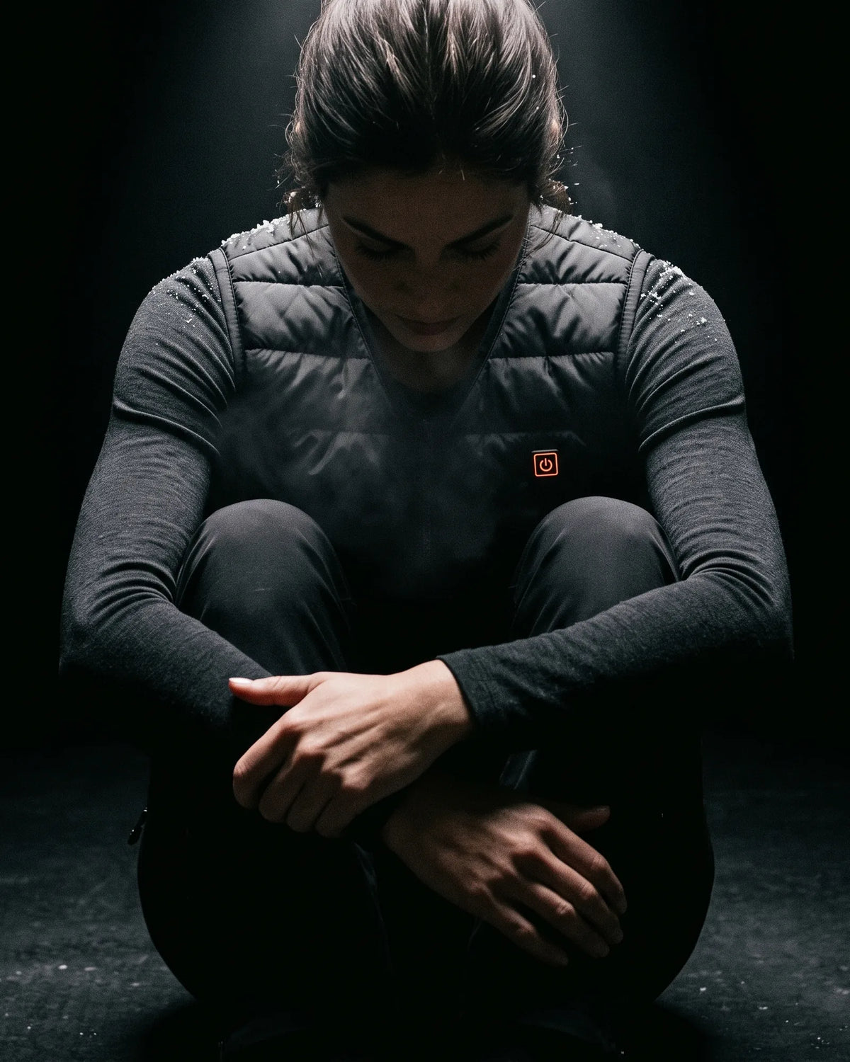 Woman in the Ridgeline heated vest sitting in the dark, lit from above, breath fogging in the cold